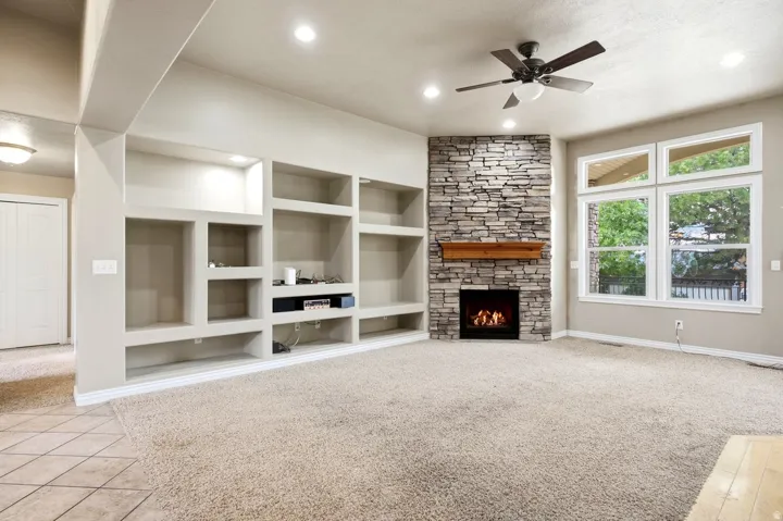 Built-in shelving, stone-clad fireplace with wood mantle, overhead lighting, ceiling fan, and multiple windows