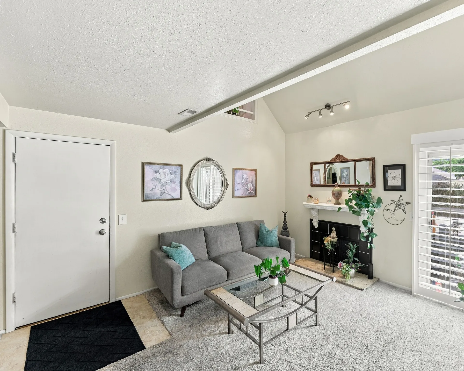 Vaulted ceiling with track lighting, fireplace with mantel, window with plantation shutters, and carpet flooring
