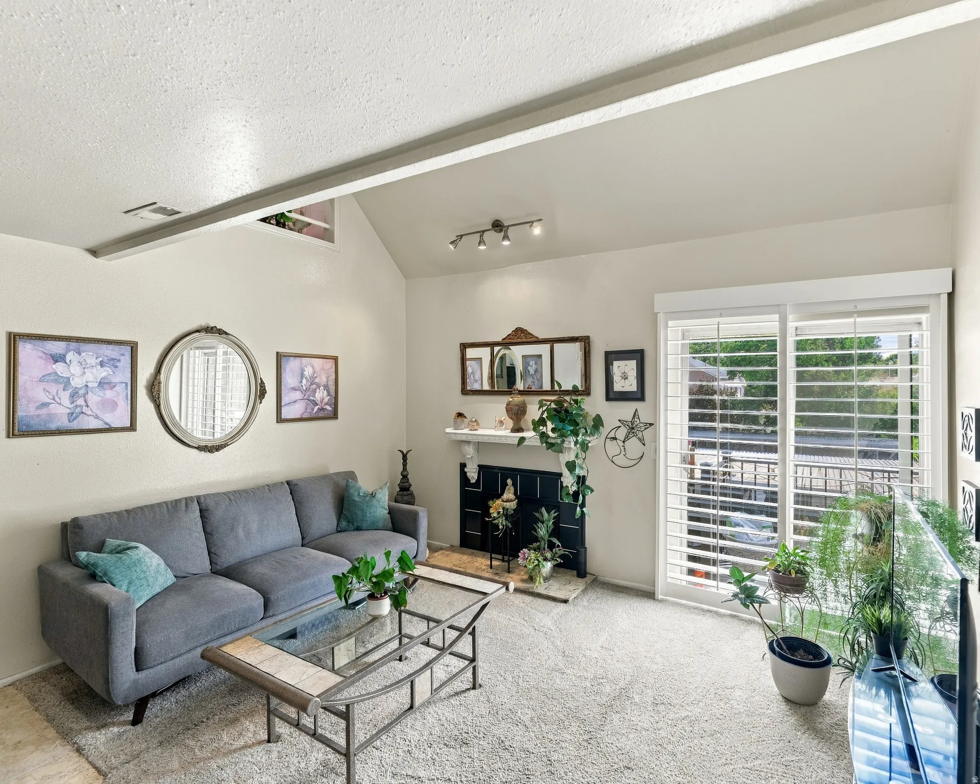 Vaulted ceiling with exposed beam, track lighting, fireplace with mantel, and sliding glass door with plantation shutters