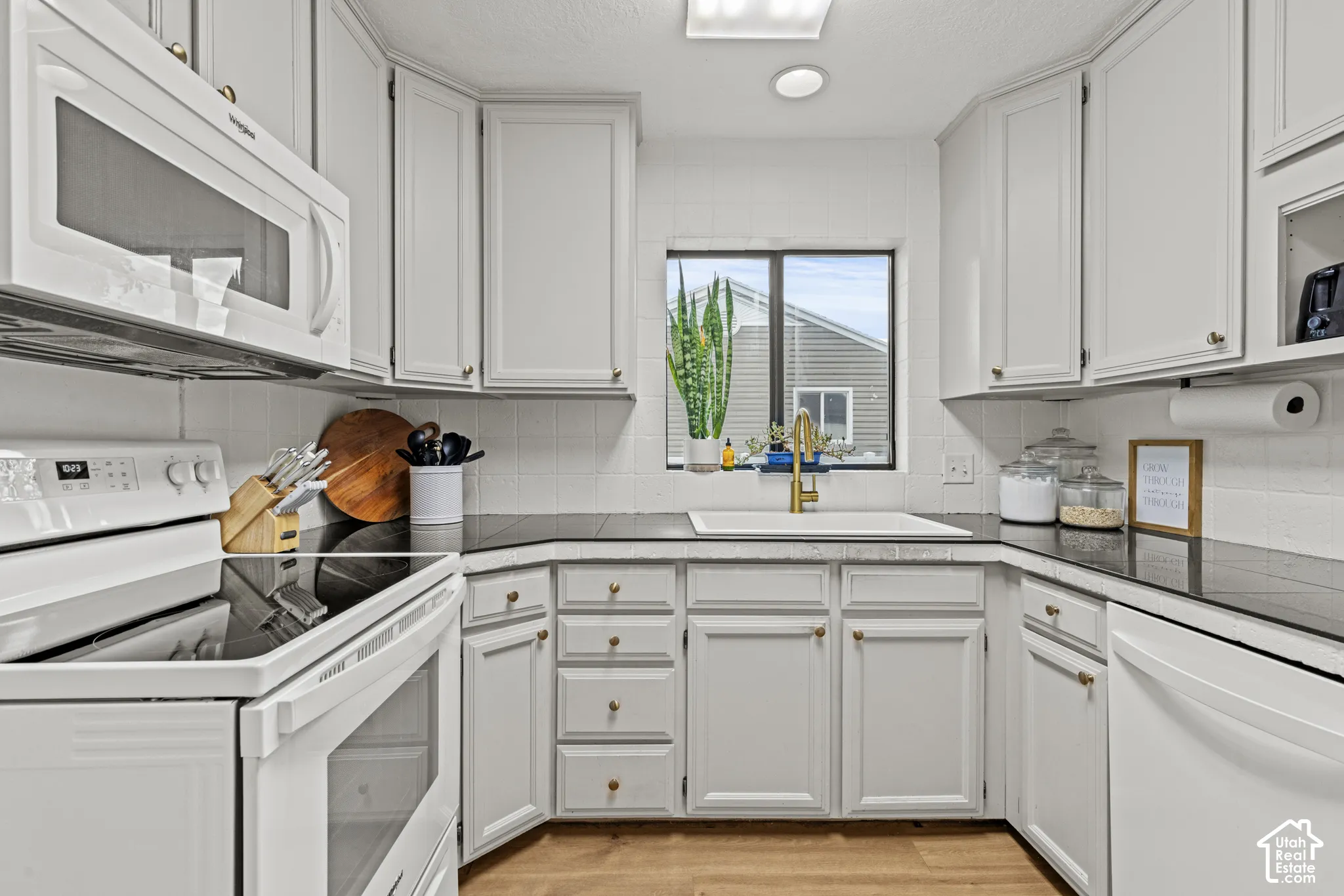 White cabinetry with gold-tone hardware, dark countertops, white tile backsplash, and wood-finish flooring