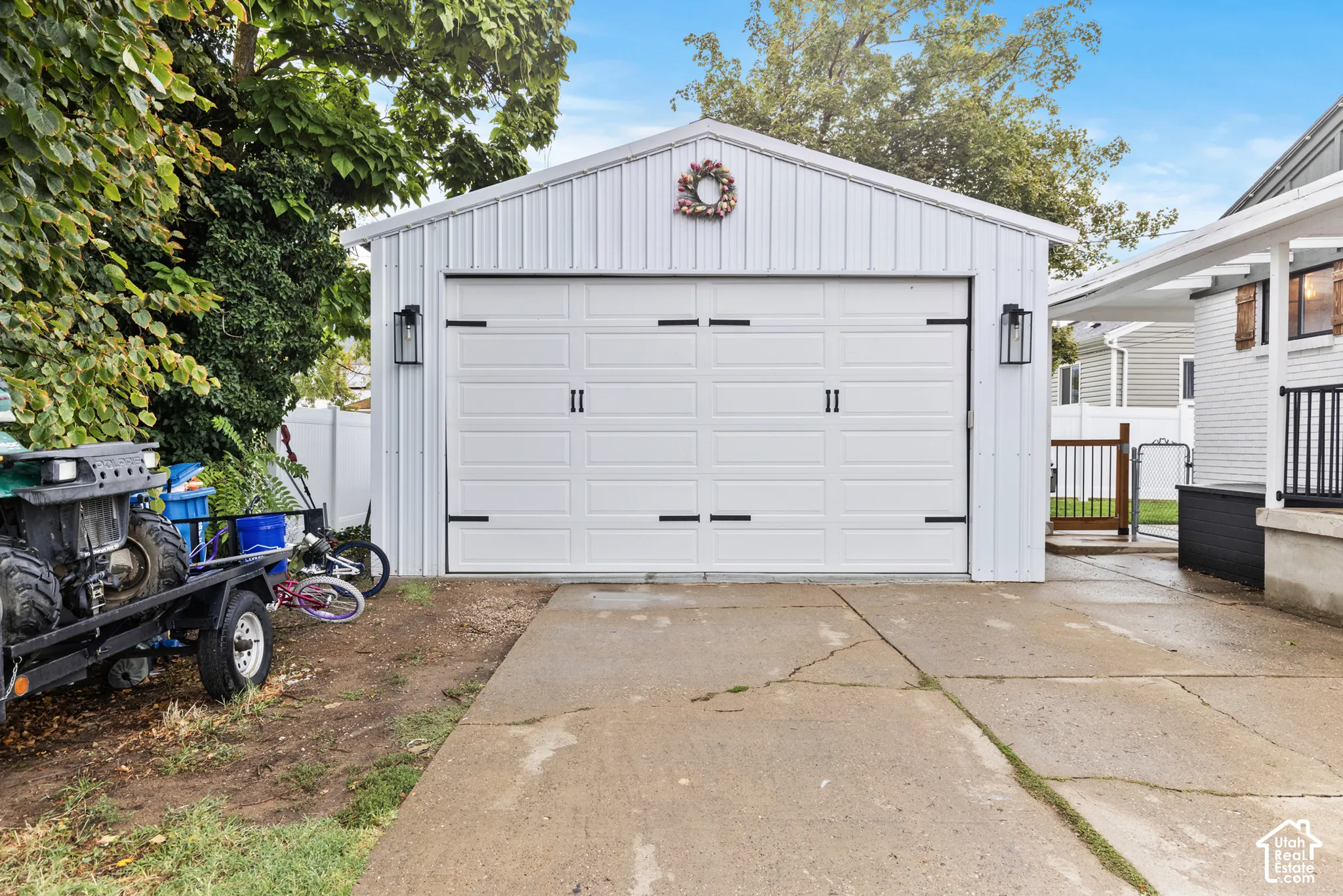 Detached garage featuring a white paneled door with black hardware, vertical siding, a gable roof, and exterior lantern lighting fixtures