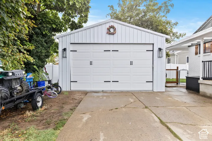 Detached garage featuring a white paneled door with black hardware, vertical siding, a gable roof, and exterior lantern lighting fixtures