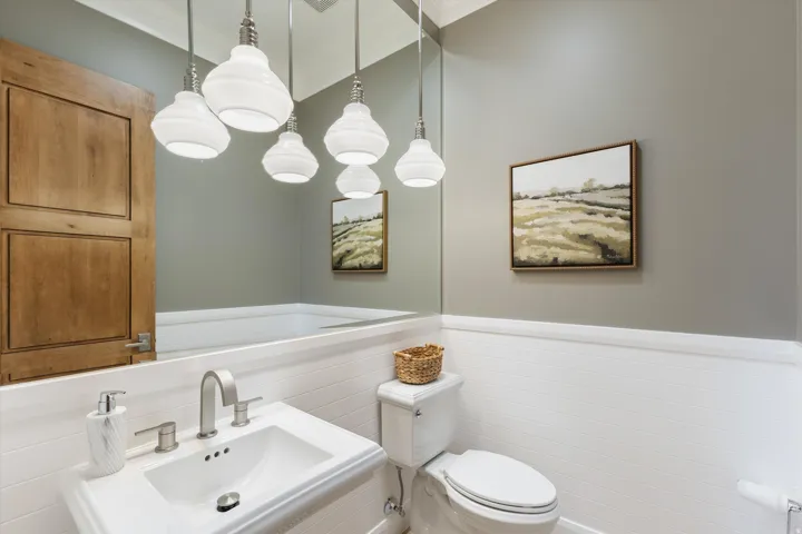 White wall paneling, light wood door, wall-mounted vanity sink, brushed nickel faucet, and multiple pendant light fixtures