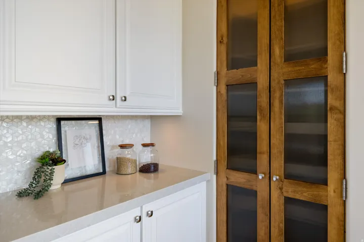 White upper and lower cabinetry, patterned tile backsplash, light-toned countertop, and wood-finish pantry doors with frosted glass panels
