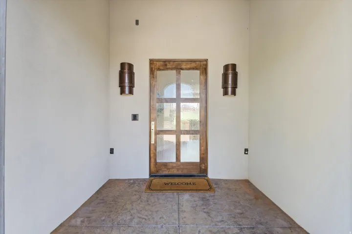 Wood-finish entry door with clear glass panels, flanked by two bronze-finish sconces, set against smooth stucco walls and textured concrete flooring