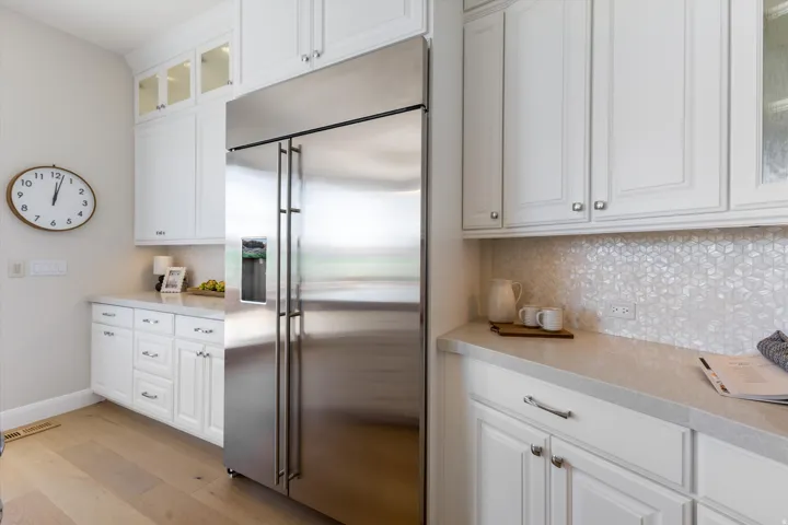 Stainless steel refrigerator, white cabinetry with silver-tone hardware, light-toned countertops, geometric mosaic tile backsplash, and wood-finish flooring