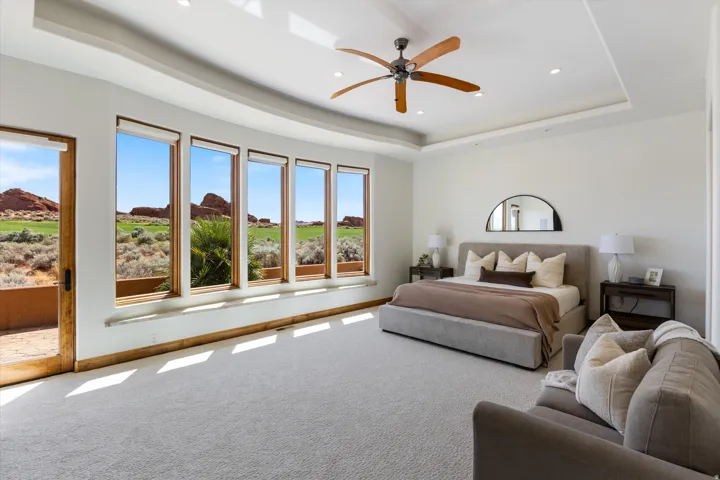 Tray ceiling with recessed lighting and a ceiling fan, light-colored carpeting, multiple large windows, glass-paneled door, and a serene outdoor backdrop