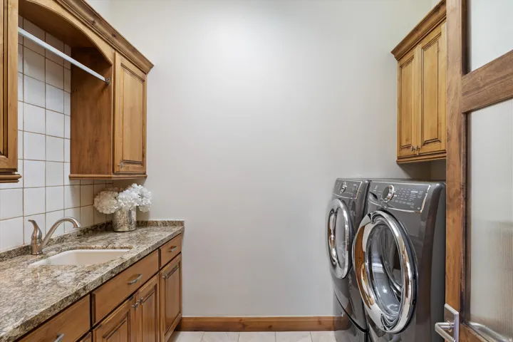Laundry area featuring wood cabinetry, stone countertops, a single basin sink with a brushed nickel faucet, and white tile backsplash