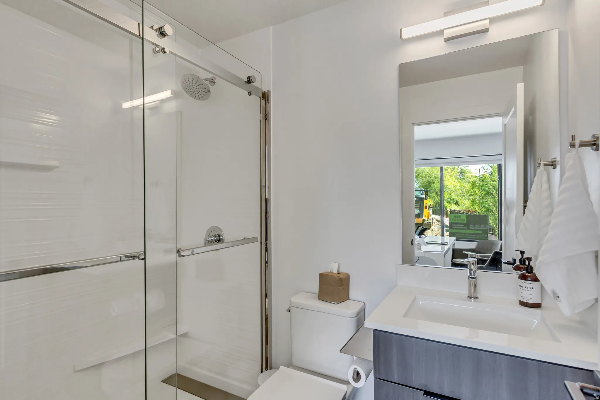 Glass-enclosed shower with a rainfall showerhead, single vanity with a white countertop and undermount sink, and a framed mirror with integrated lighting