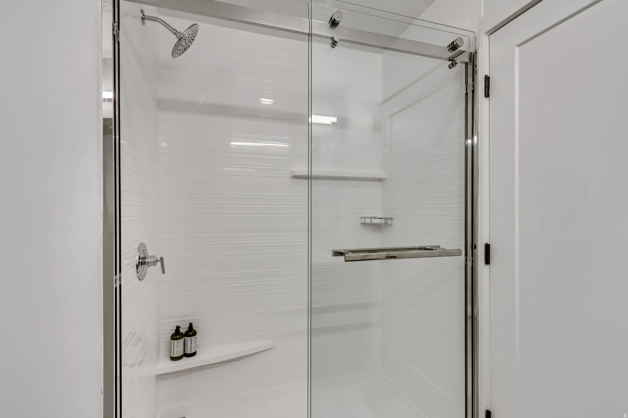 Glass-enclosed shower featuring a rainfall showerhead, white textured tile, a built-in corner shelf, and polished chrome fixtures