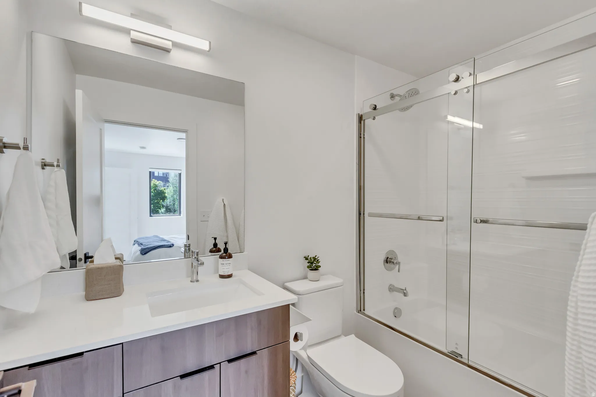 Bathroom featuring a floating wood-finish vanity with an integrated rectangular sink, a frameless mirror, and a shower-tub combination with sliding glass doors
