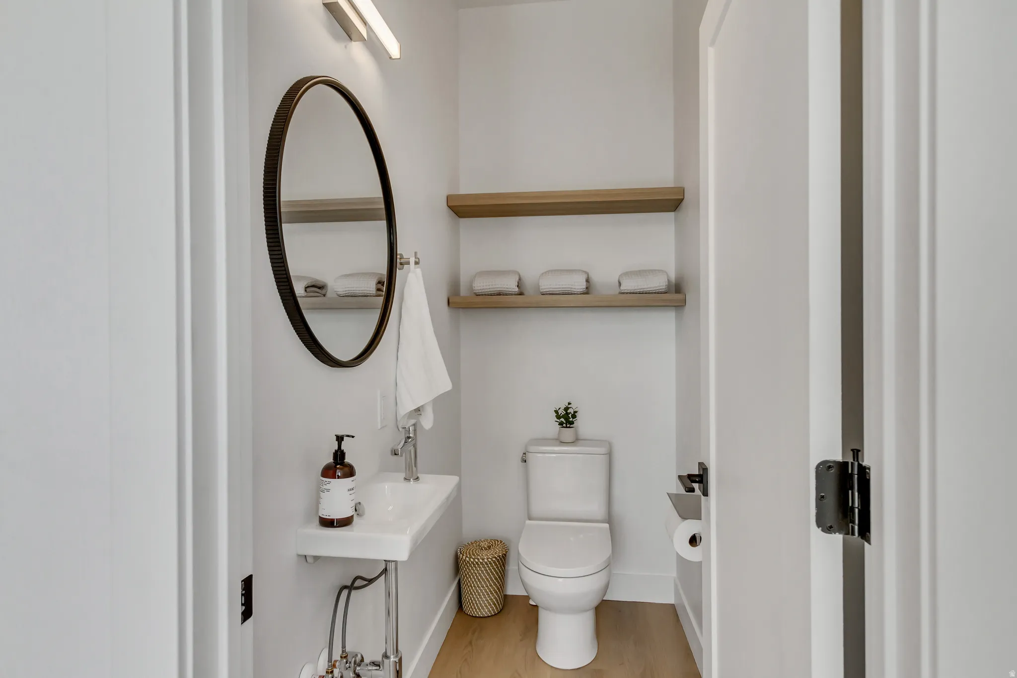 Wall-mounted ceramic sink, round framed mirror, wood-finish flooring, and floating wood shelves