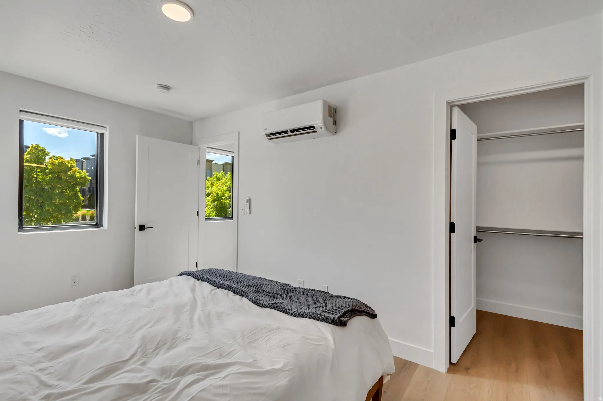 Recessed ceiling lighting, mini-split air conditioning unit, light wood-finish flooring, two windows, and an open closet with shelving and hanging rods