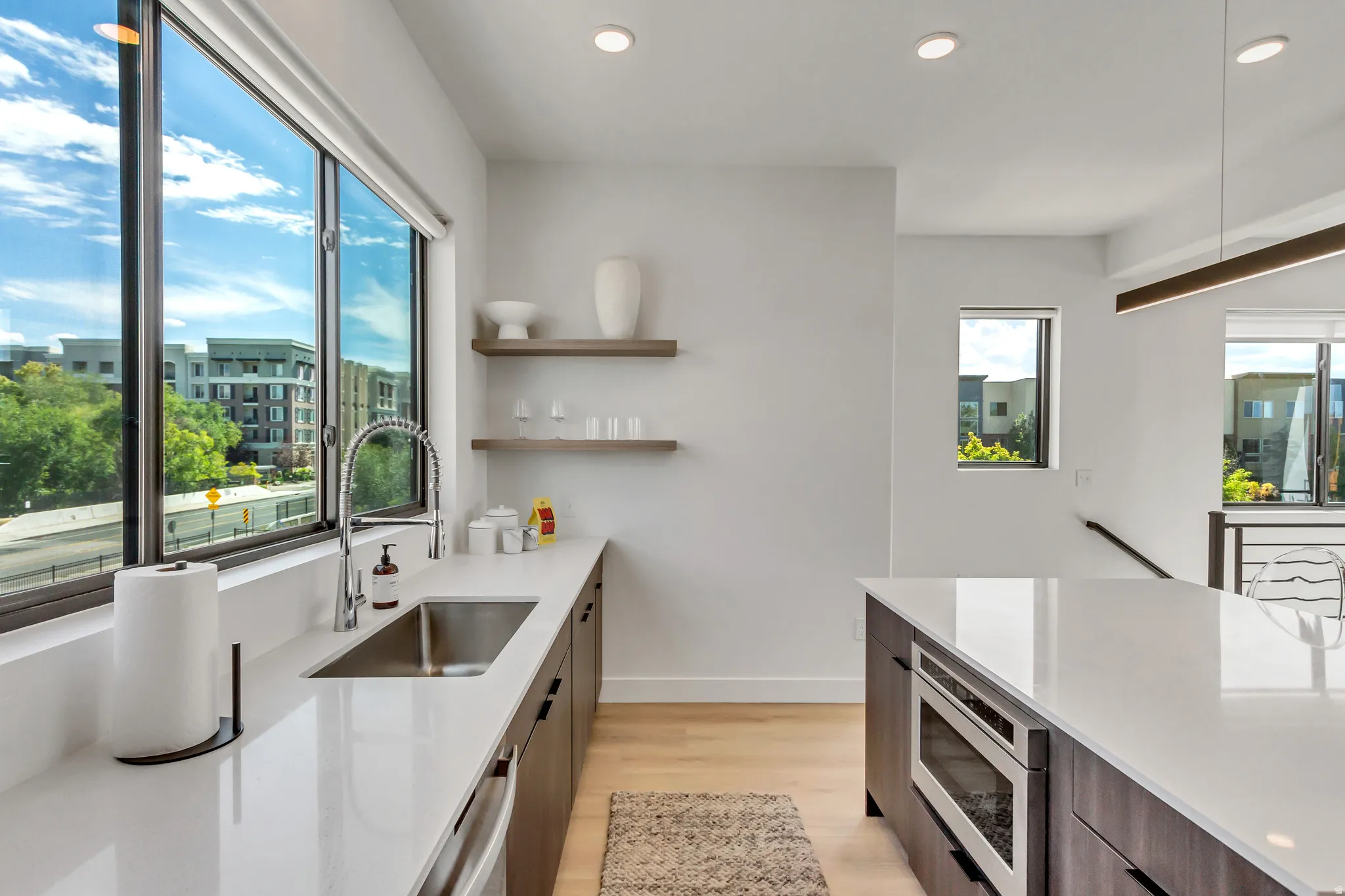 Modern kitchen featuring expansive windows, light wood-finish flooring, white countertops, dark wood cabinetry, and an integrated microwave