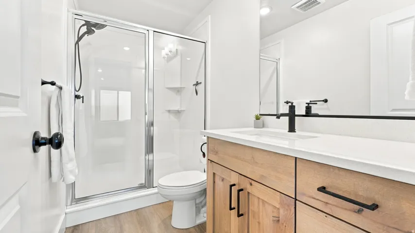 Glass-enclosed shower with matte black fixtures, wood-finish flooring, light wood vanity with matte black hardware, white countertop, and integrated sink