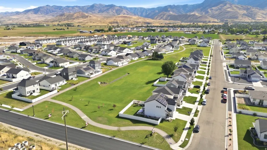 Expansive residential community featuring modern homes with dark roofs and light-colored siding, a large central green space, paved pathways, and mountain scenery