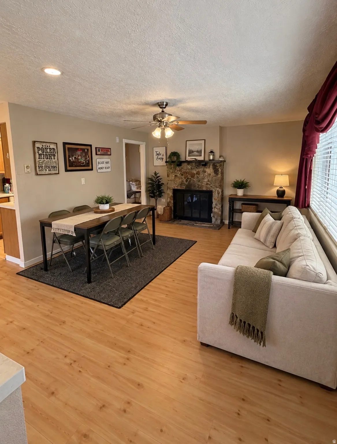 Wood-finish flooring throughout, stone-faced fireplace with a black insert, ceiling fan with integrated lighting, recessed lighting, and horizontal blinds