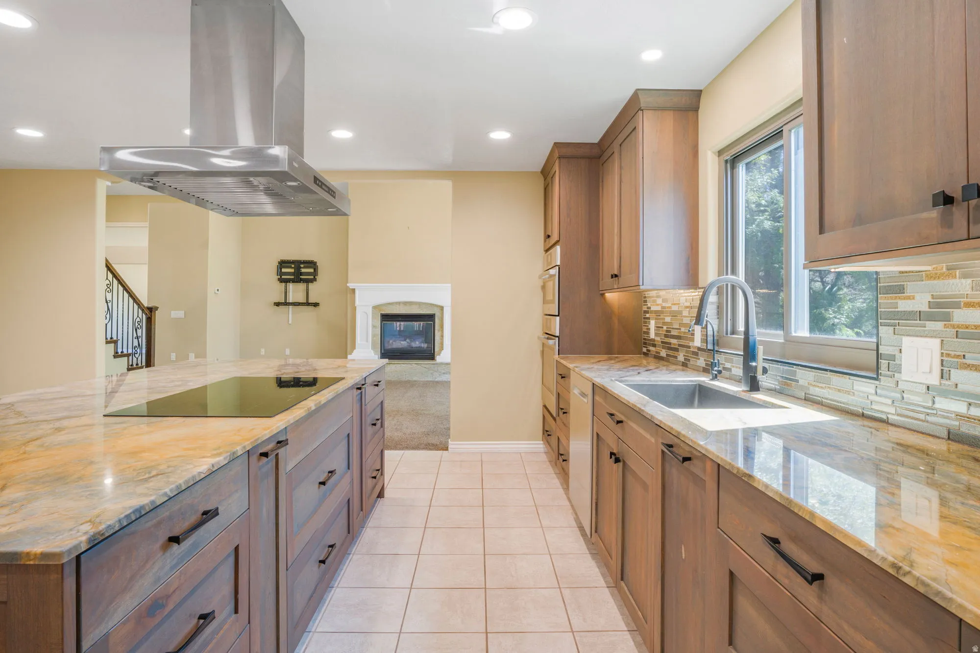 Kitchen featuring a large island with an integrated cooktop, stainless steel range hood, stone countertops, wood-finish cabinetry, and a tile backsplash