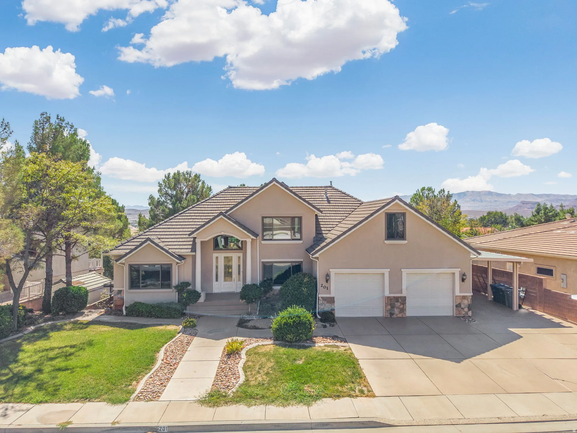 Stucco exterior with a tiled roof, mature landscaping, a two-car garage, and a covered carport