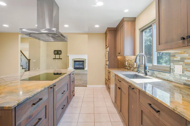 Kitchen featuring a large island with an integrated cooktop, stainless steel range hood, stone countertops, wood-finish cabinetry, and a tile backsplash