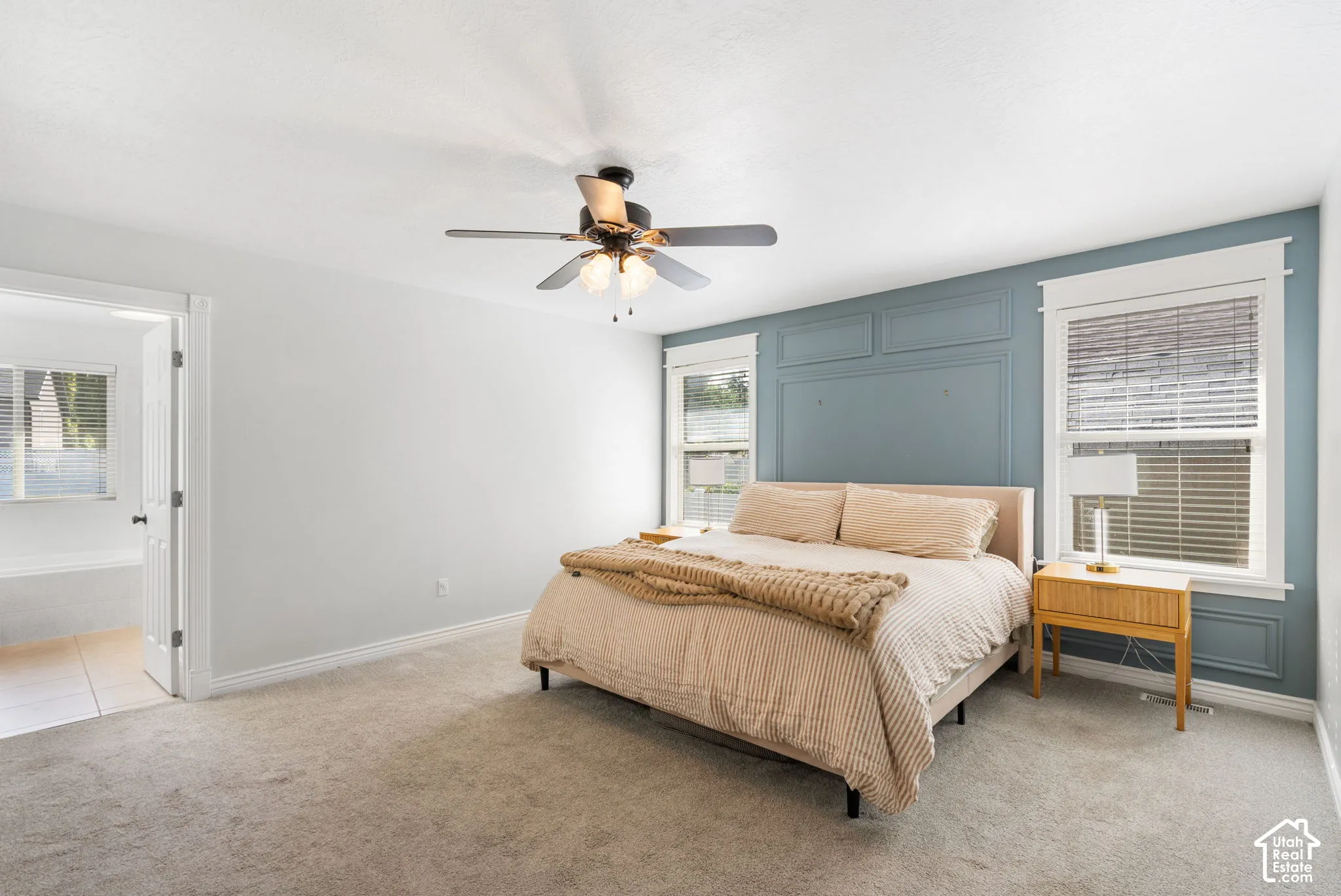 Carpeted bedroom featuring a ceiling fan, accent wall with panel molding, and two windows with blinds