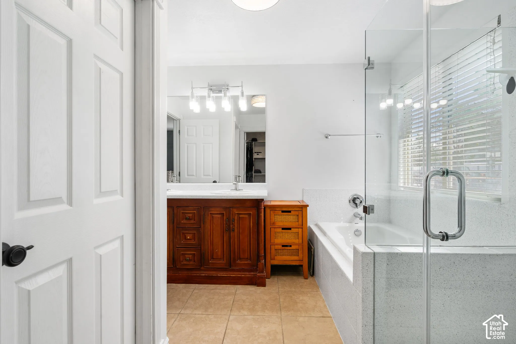 Bathroom featuring a wood-finish vanity with dual sinks, a large mirror with integrated lighting, a glass-enclosed shower, a built-in tub, and tile flooring