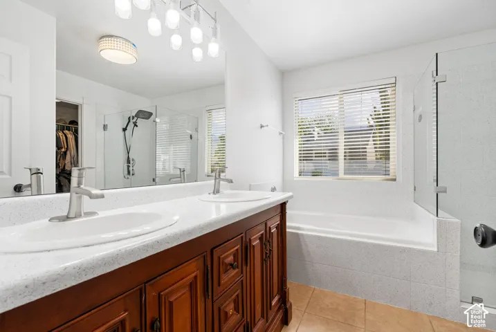 Dual sink vanity with wood cabinetry, large soaking tub, glass-enclosed shower, and tile flooring