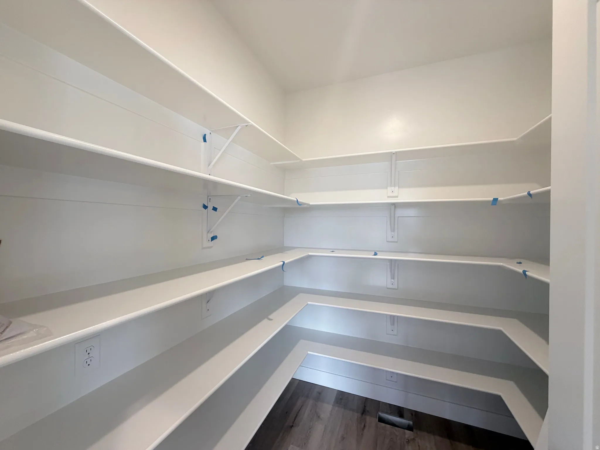 Walk-in pantry featuring multiple white shelves, wood-finish flooring, and electrical outlets
