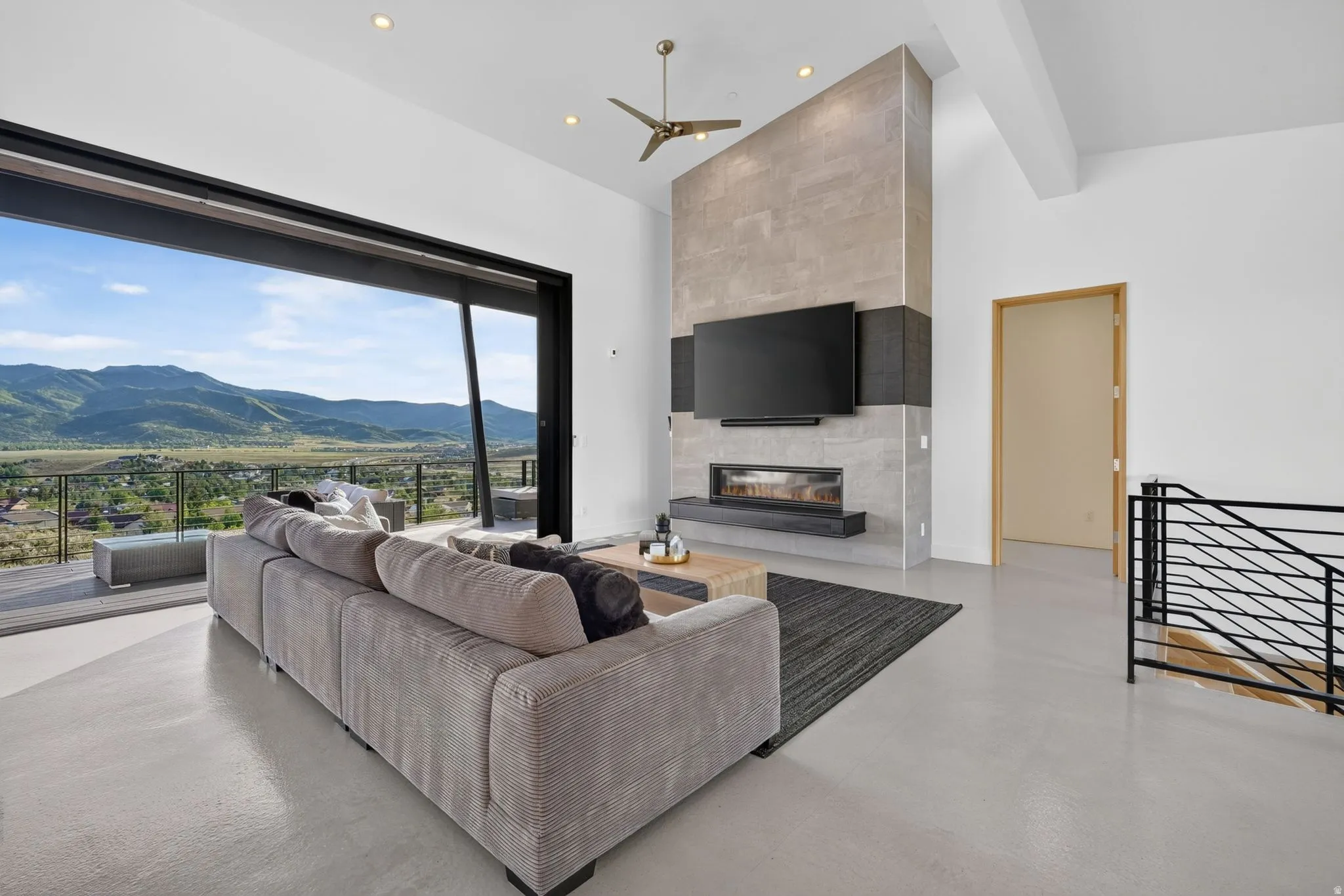 Expansive floor-to-ceiling sliding glass doors, a prominent fireplace with a built-in electric firebox, a high ceiling with a ceiling fan, and sleek metal stair railing