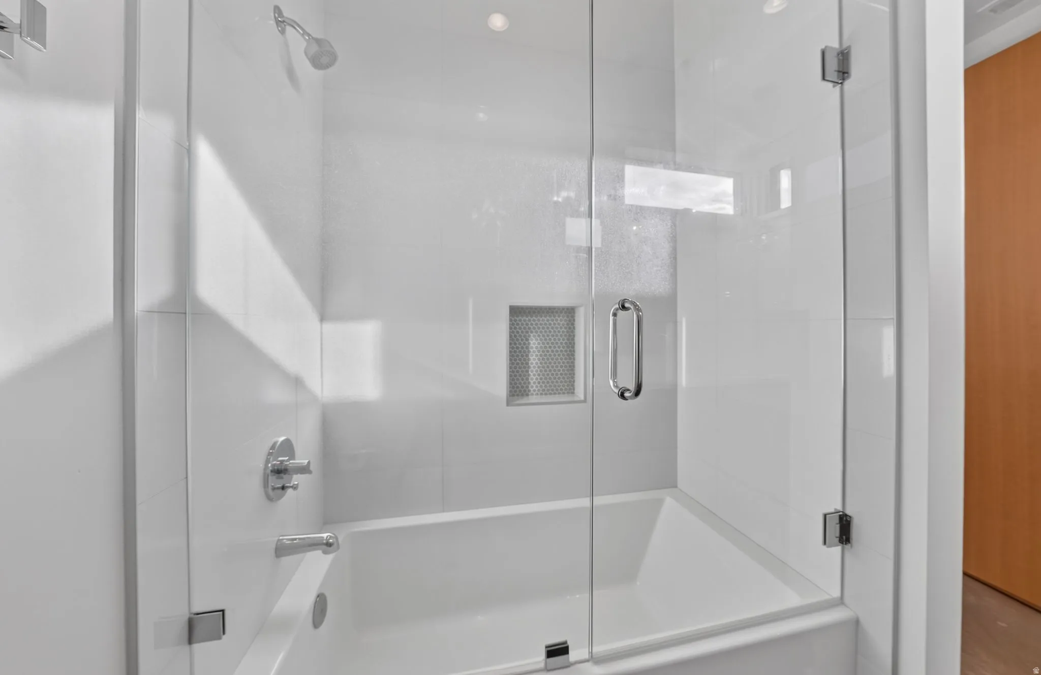 Bathtub with a glass shower enclosure, white tiled surround, built-in shower niche, and chrome fixtures