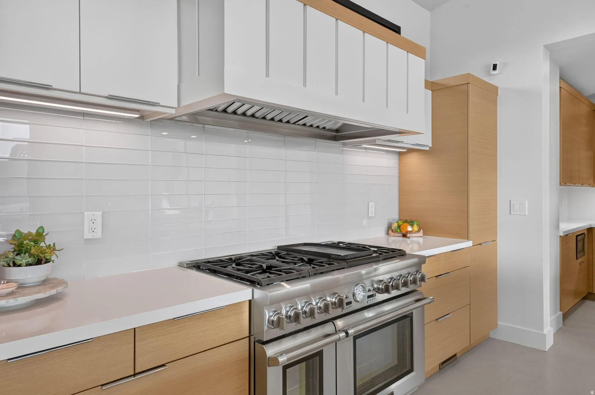 Professional-grade stainless steel range with double ovens, white subway tile backsplash, light wood-finish lower cabinetry, white upper cabinetry, and white countertops