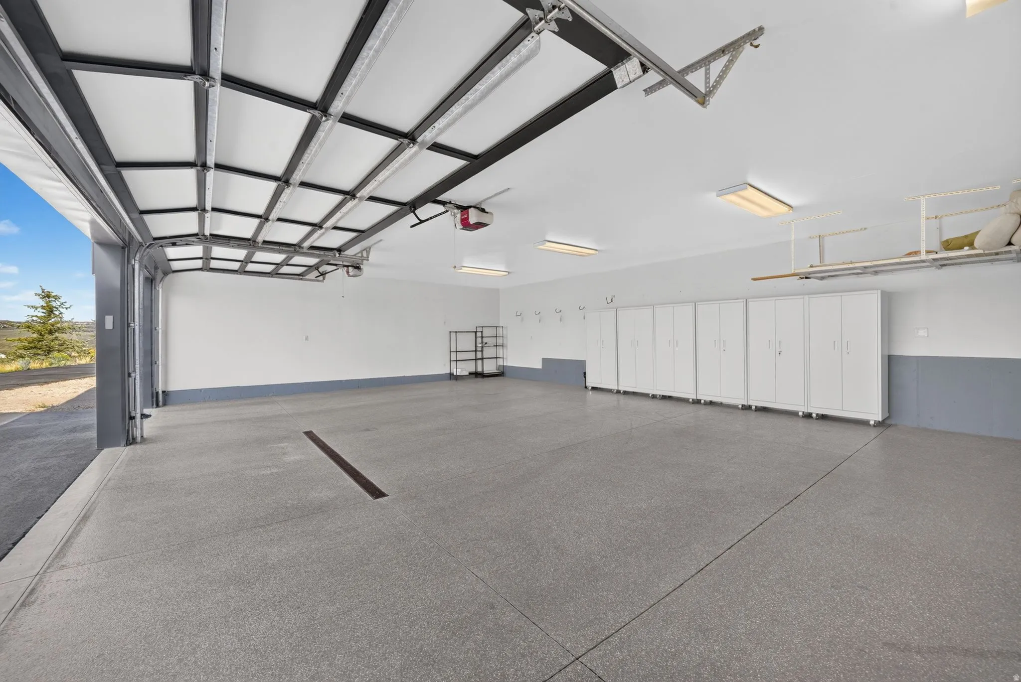 Garage with an open overhead door, overhead lighting, an epoxy-coated floor, a row of built-in storage cabinets, and overhead storage racks