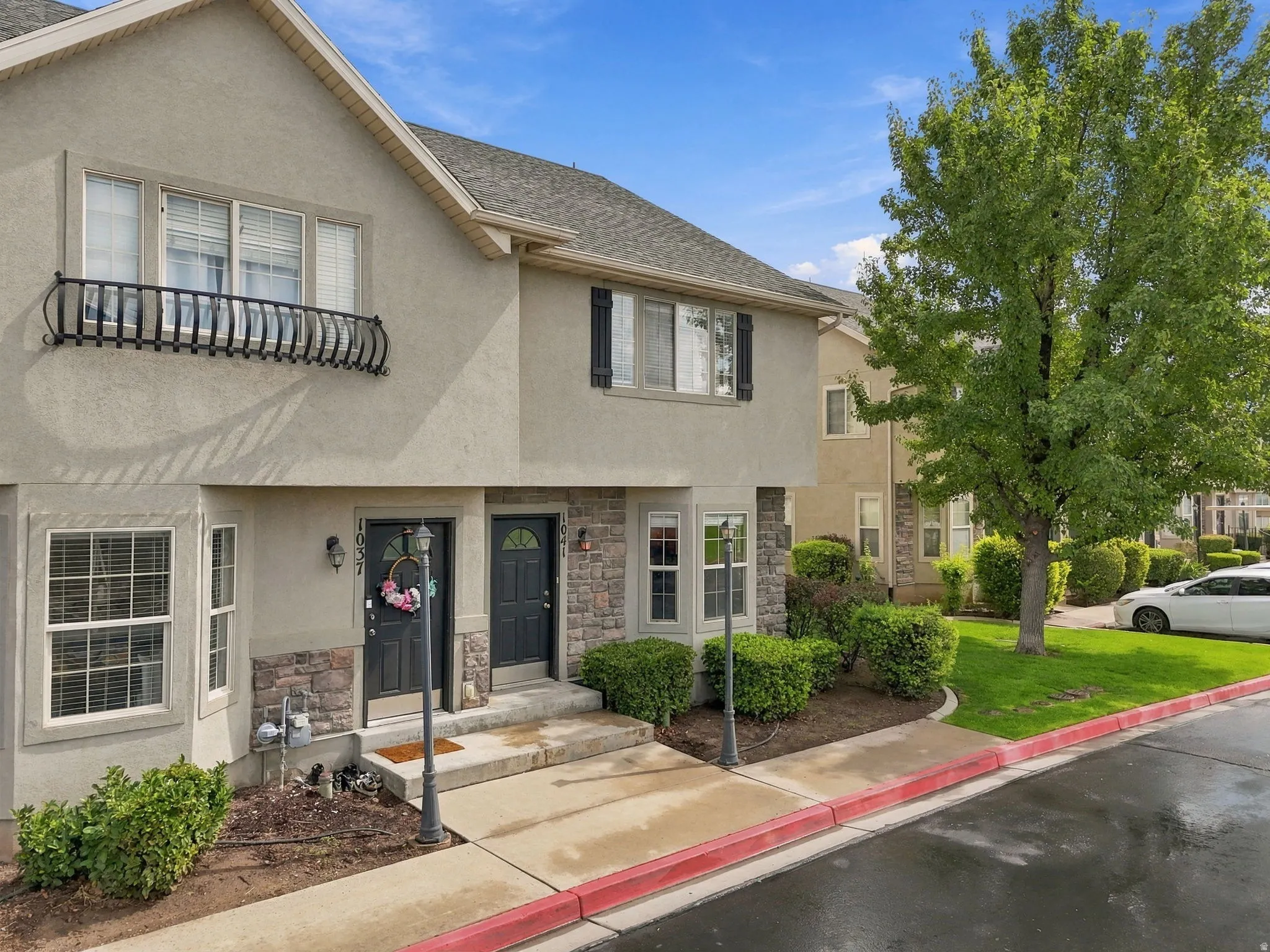 Stucco exterior with stone veneer accents, individual entryways, decorative iron balcony railing, and mature landscaping