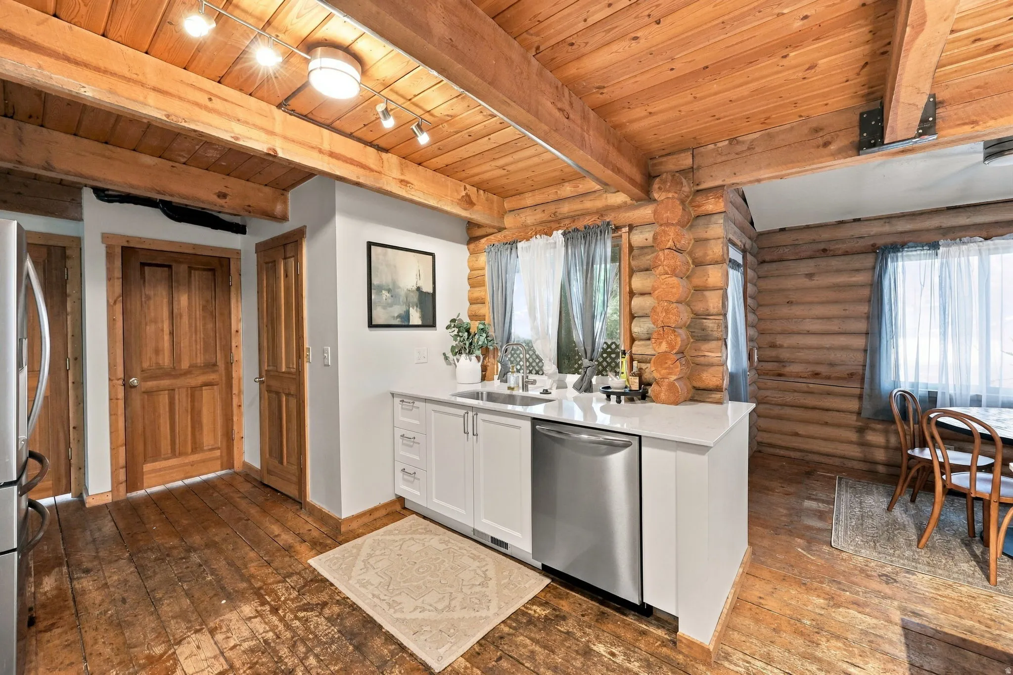 Exposed wood beam ceiling, wood-finish flooring, white cabinetry, integrated stainless steel dishwasher, and log wall construction