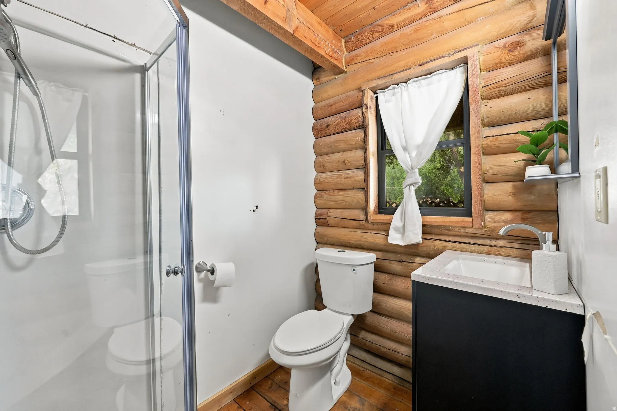 Log wall paneling, wood-finish flooring, glass-enclosed shower, white ceramic toilet, and a vanity with a white countertop