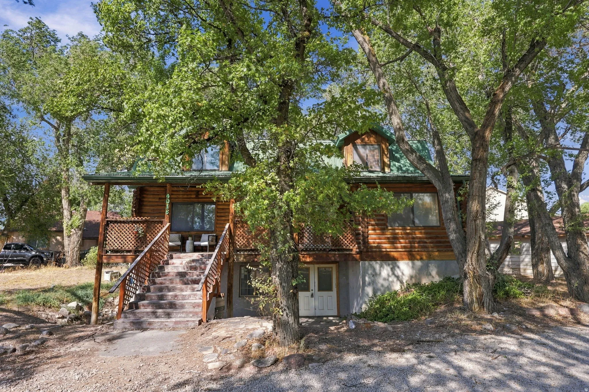 Log cabin style exterior with a green metal roof, multiple mature trees, and an upper-level deck with lattice railing