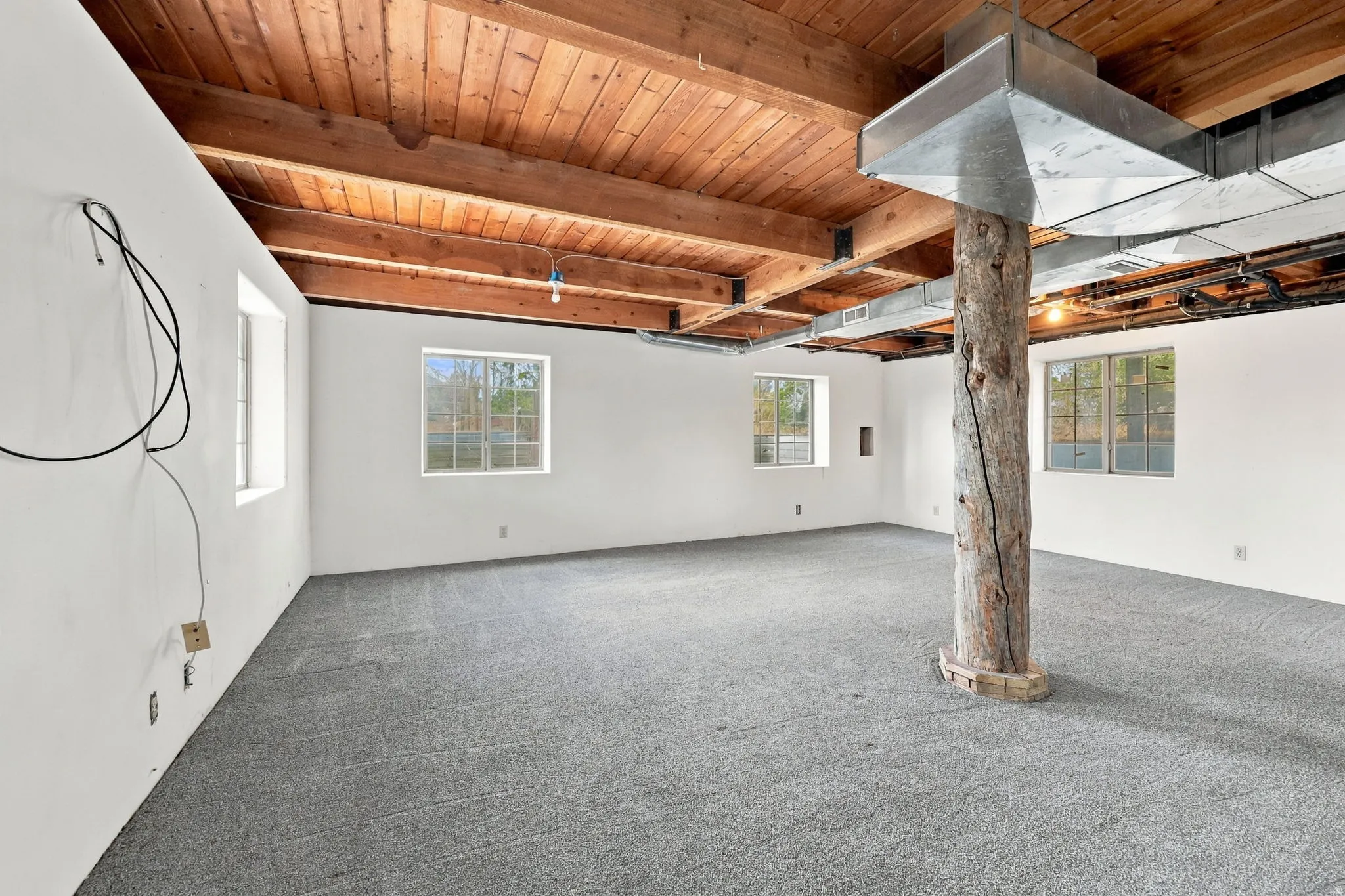 Exposed wood ceiling beams, white painted walls, gray carpeting, a natural wood support column, and multiple windows
