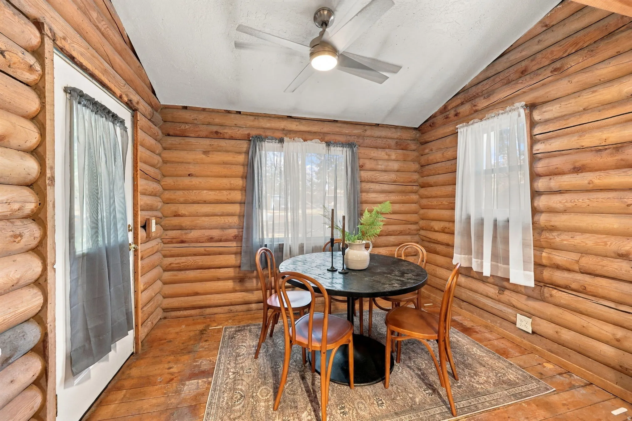 Log wall construction, wood-finish flooring, white ceiling, ceiling fan with integrated lighting, and a white door with glass insert