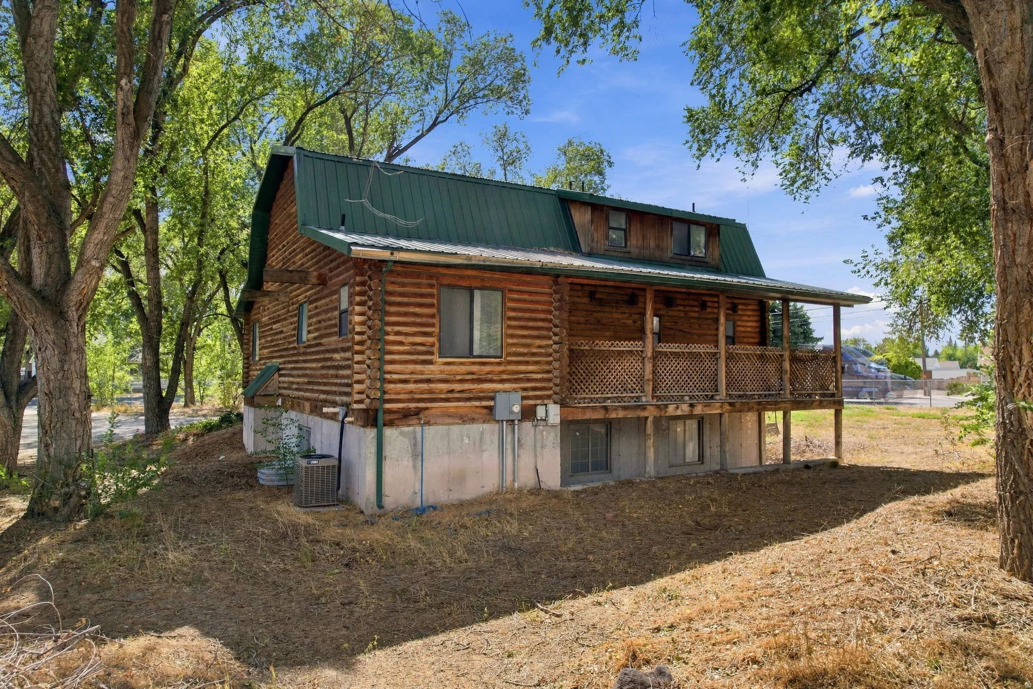 Log cabin exterior with a green gambrel roof, featuring a covered porch with a lattice railing and a concrete foundation