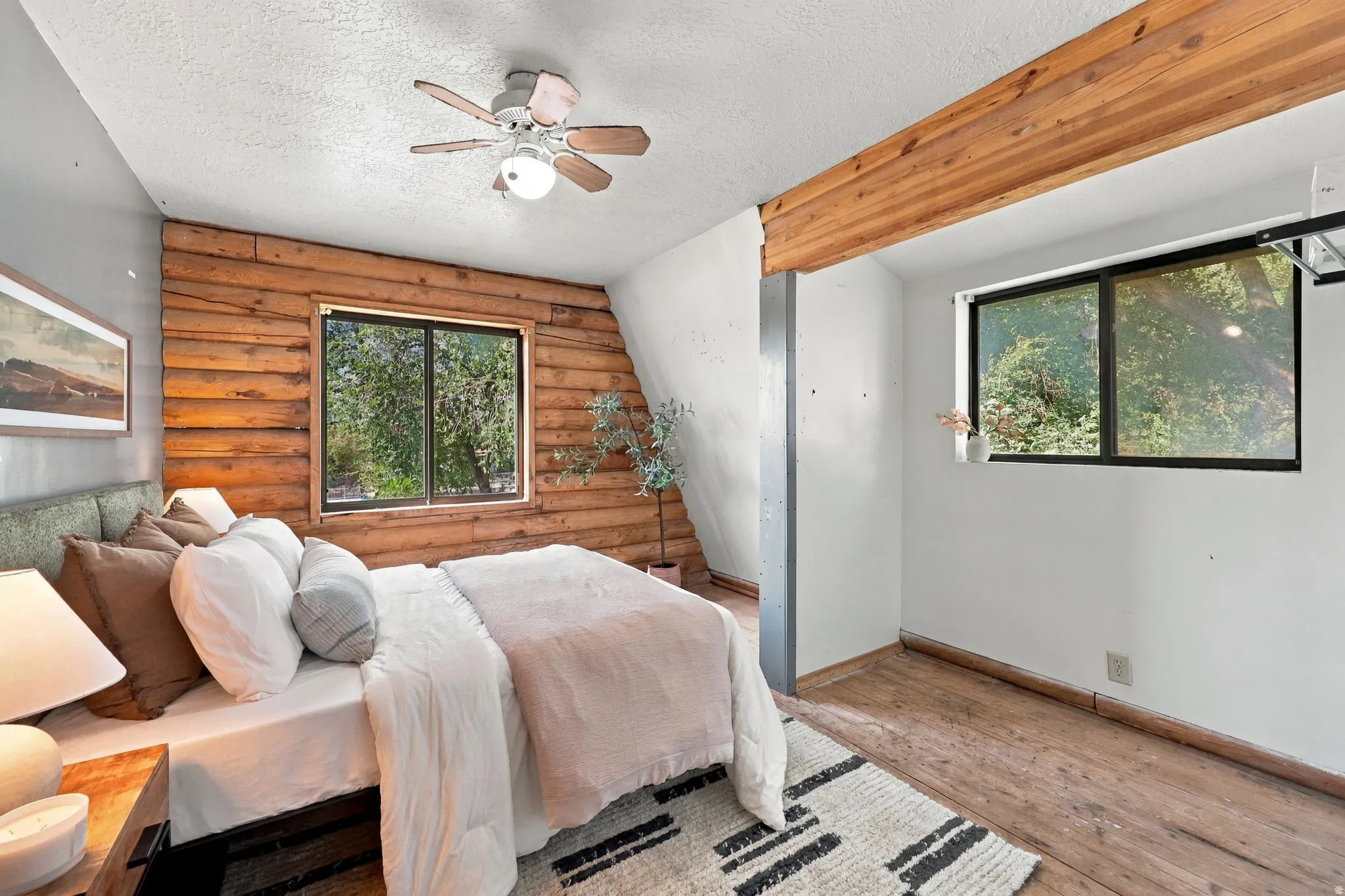 Wood-paneled accent wall, exposed wood beam, wood-finish flooring, two large windows, and a ceiling fan with integrated lighting