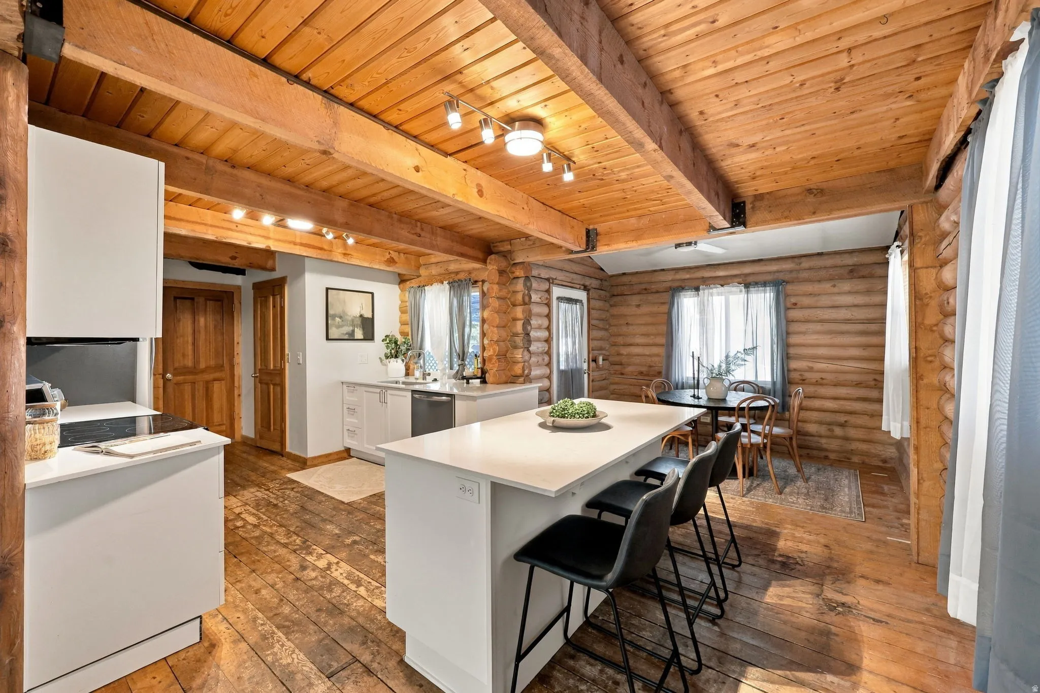 Exposed wood beam ceiling, wood-finish flooring, white cabinetry, stainless steel dishwasher, and a white countertop island with integrated seating