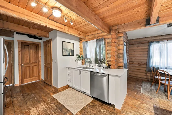 Exposed wood beam ceiling, wood-finish flooring, white cabinetry, integrated stainless steel dishwasher, and log wall construction