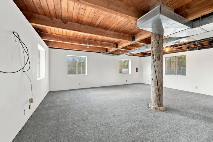 Exposed wood ceiling beams, white painted walls, gray carpeting, a natural wood support column, and multiple windows