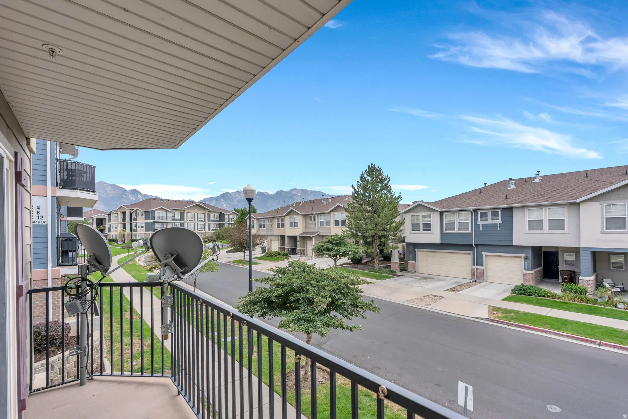 Balcony with black metal railing, overlooking a paved road with sidewalks, green landscaping, and distant mountains