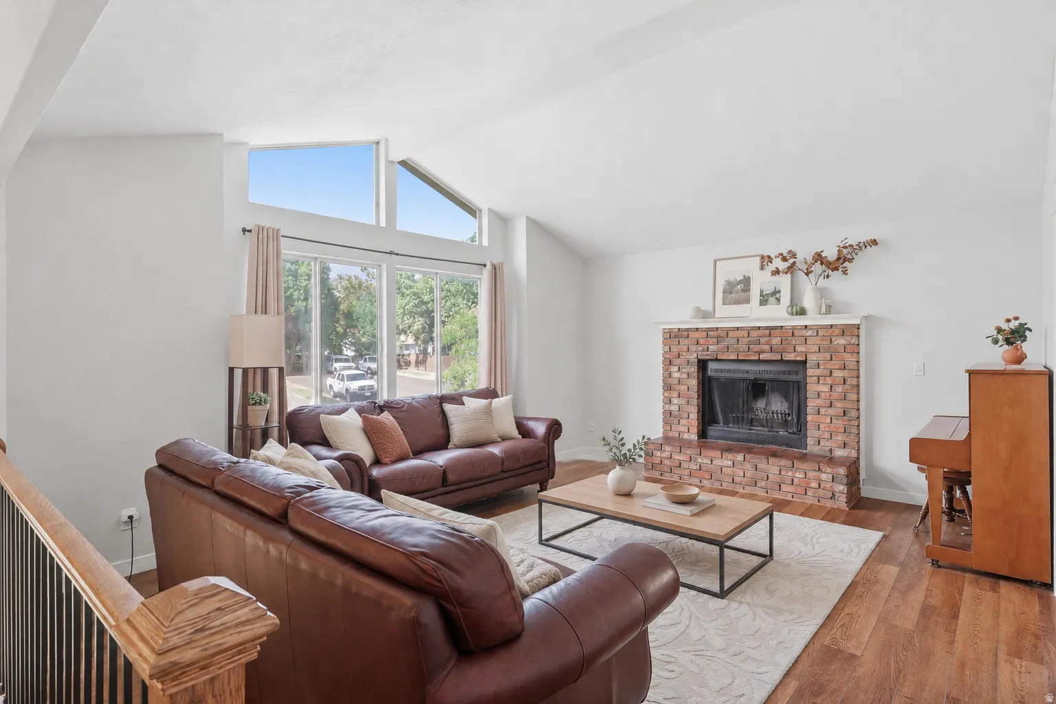 Vaulted ceiling, brick-surround fireplace, large windows, wood-finish flooring, and a decorative wood stair railing
