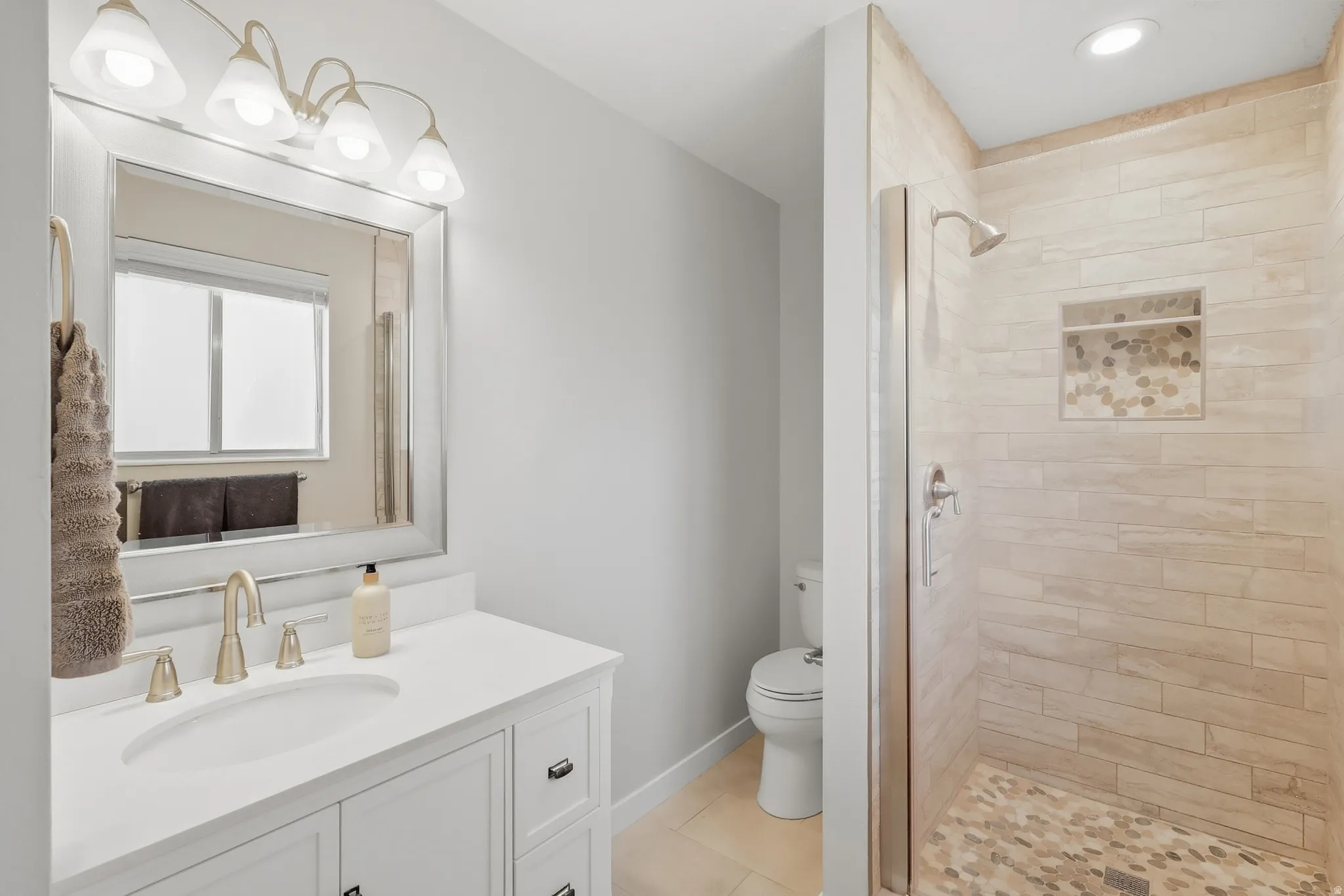 White vanity with an integrated sink and brushed nickel faucet, large framed mirror, and a tiled shower with a niche and pebble-tile flooring