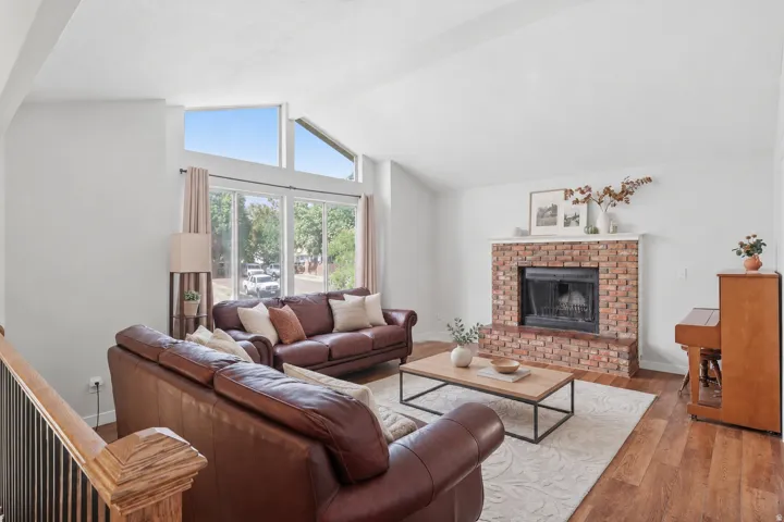 Vaulted ceiling, brick-surround fireplace, large windows, wood-finish flooring, and a decorative wood stair railing