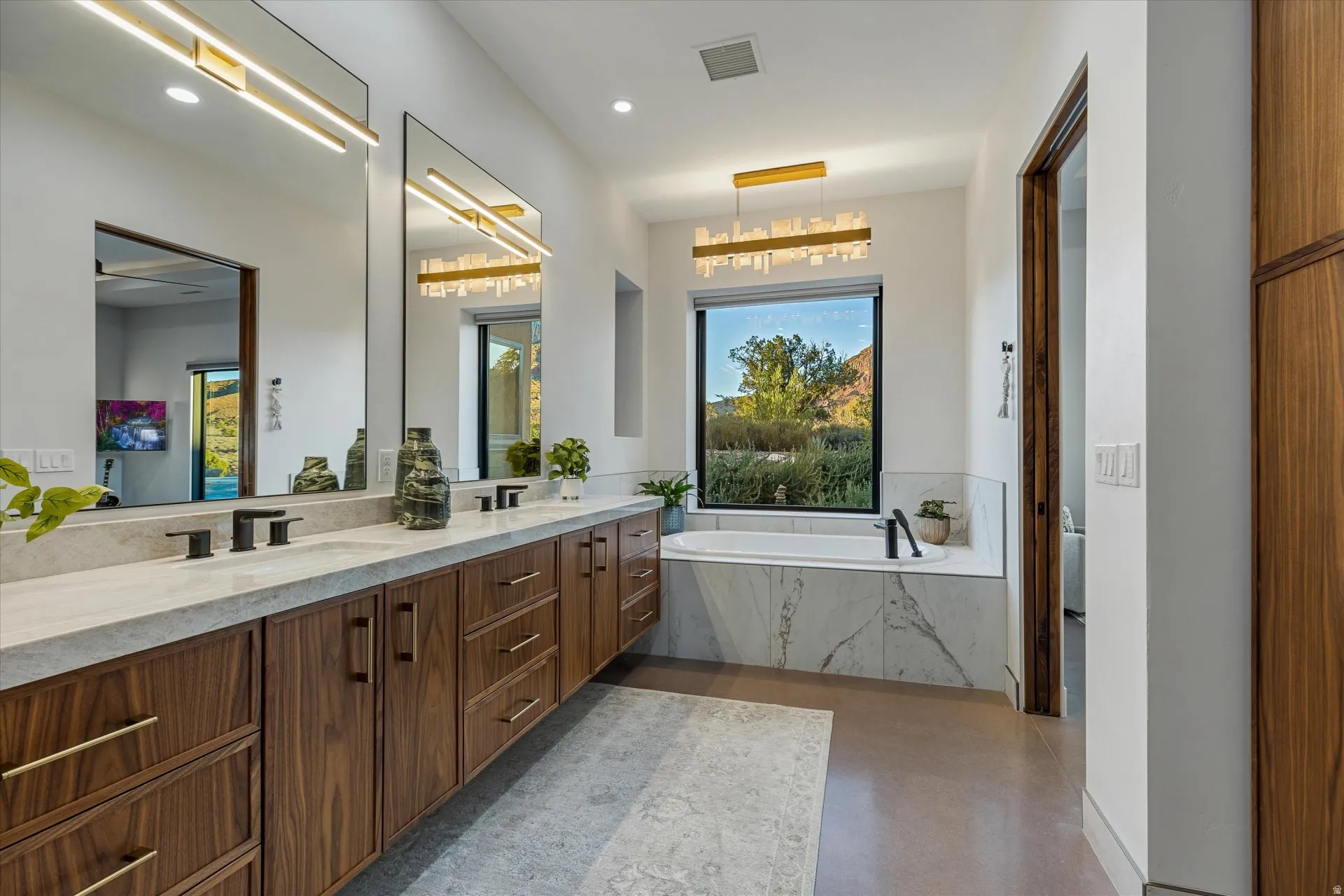 Primary bath soaking tub beneath an alabaster-style chandelier, framed by a picture window looking out at Mt. Kinesava, plus a floating dual vanity.