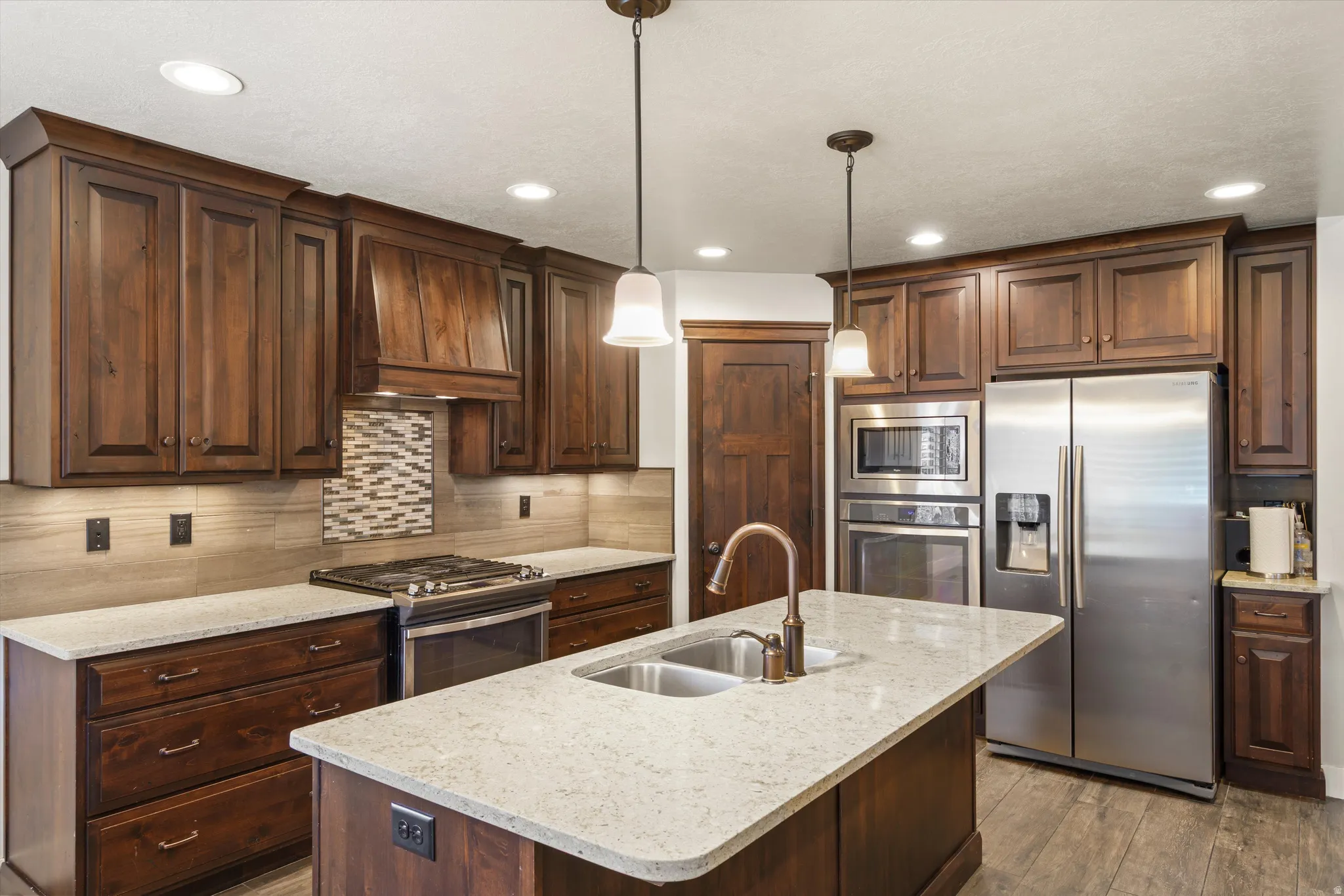 Kitchen featuring wood-finish cabinetry, light-toned countertops, stainless steel appliances, a center island with sink, and recessed lighting