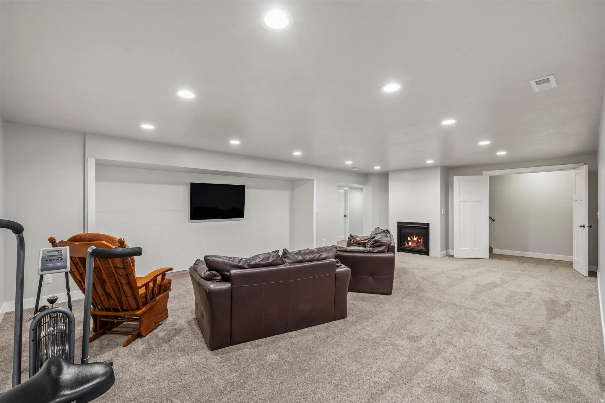 Recessed lighting illuminates a carpeted living space with a wall-mounted television, built-in fireplace, and paneled doors